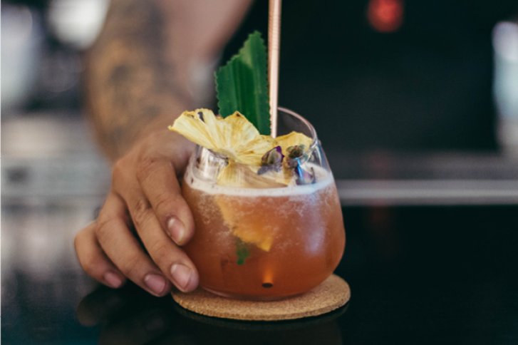 Ein Cocktail mit Ananas und Minze wird von einer Hand auf einem Tresen serviert.