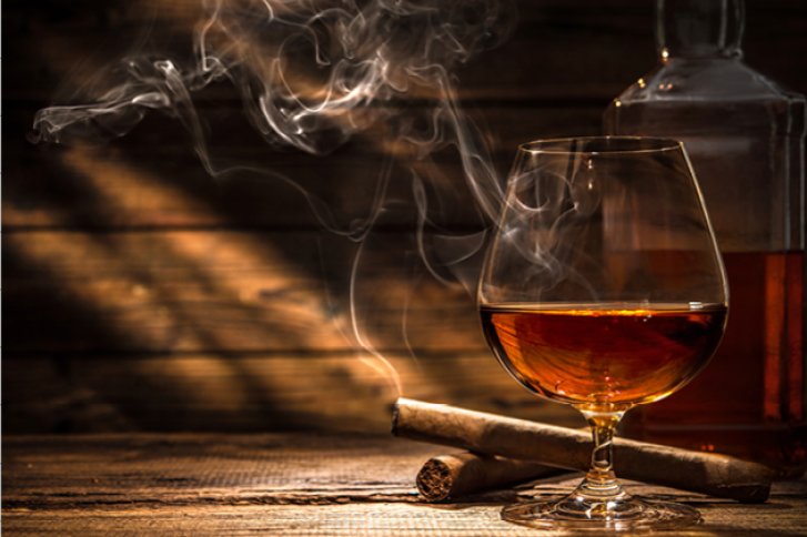 Ein Cognacglas steht neben einer qualmenden Zigarre auf einem Holztisch.