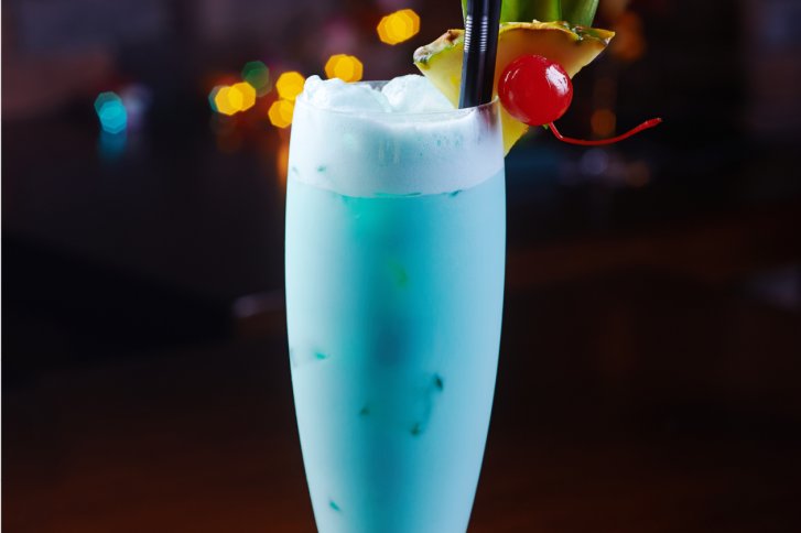 Hohes Glas mit blauem Getränk, verziert mit Ananas, Kirsche, Strohhalm; unscharfer Hintergrund.