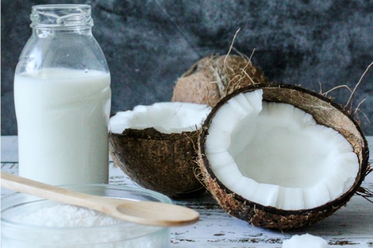 Kokosnüsse und Glasflasche stehen auf Holztisch, daneben Holzlöffel und Schale mit Kokosraspeln.