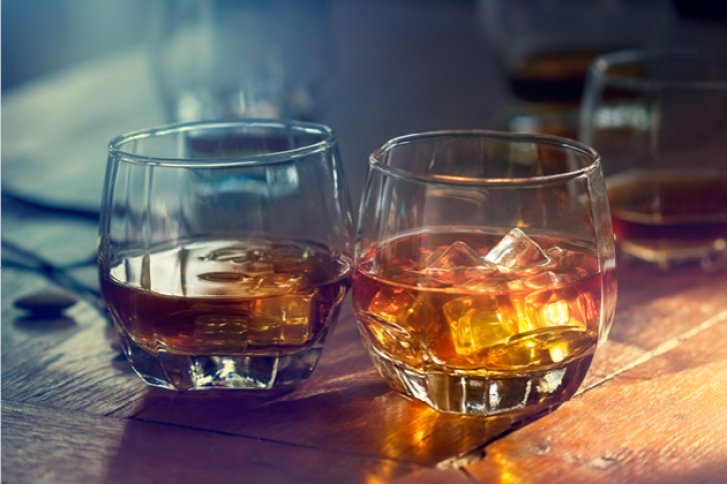 Zwei Whiskygläser stehen auf einem Holztisch, eines mit Eiswürfeln und Sonnenlicht darauf.