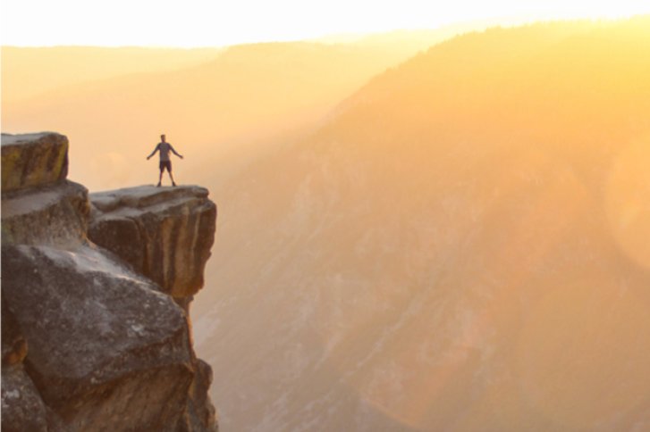 Eine Person steht mit ausgebreiteten Armen auf einem Felsvorsprung bei Sonnenuntergang.