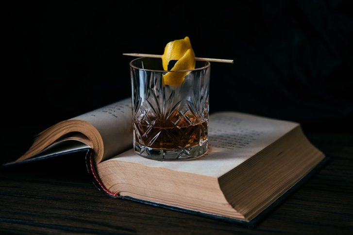 Kristallglas mit gelbem Zitrusdreh auf Holzspieß steht auf aufgeschlagenem Buch, dunkler Hintergrund.