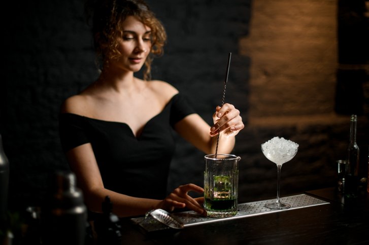 Frau rührt grünes Getränk in Glas neben Cocktailglas mit Eis in dunkler Bar.