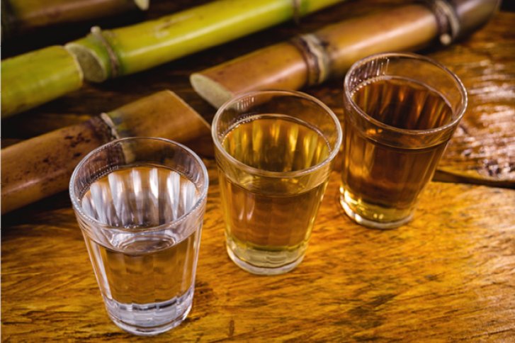 Drei Gläser mit farbigen Flüssigkeiten stehen auf Holz, umgeben von geschnittenen Zuckerrohrstangen.