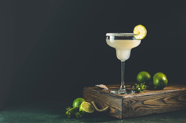 Glas Margarita mit Limettenscheibe steht auf Holzkiste, daneben liegen ganze und geschnittene Limetten.