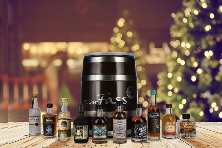Verschiedene Miniatur-Flaschen und ein Fass stehen auf einem Holztisch. Hintergrund: verschwommene Weihnachtsbeleuchtung.