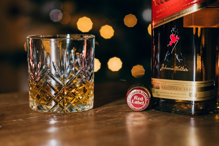 Kristallglas mit Whisky, stehend neben geöffneter Whiskyflasche, beleuchtet vor unscharfen, goldenen Lichtern.