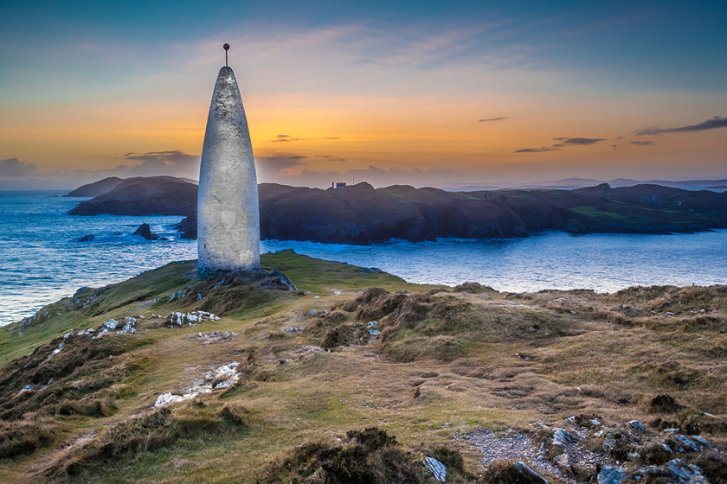 Steinmonument steht auf grasbedecktem Hügel, umgeben von Meer und Inseln im Sonnenuntergang.