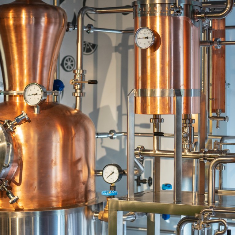 Un distillateur en cuivre se trouve dans une brasserie, entouré de tuyaux et d'appareils de mesure.