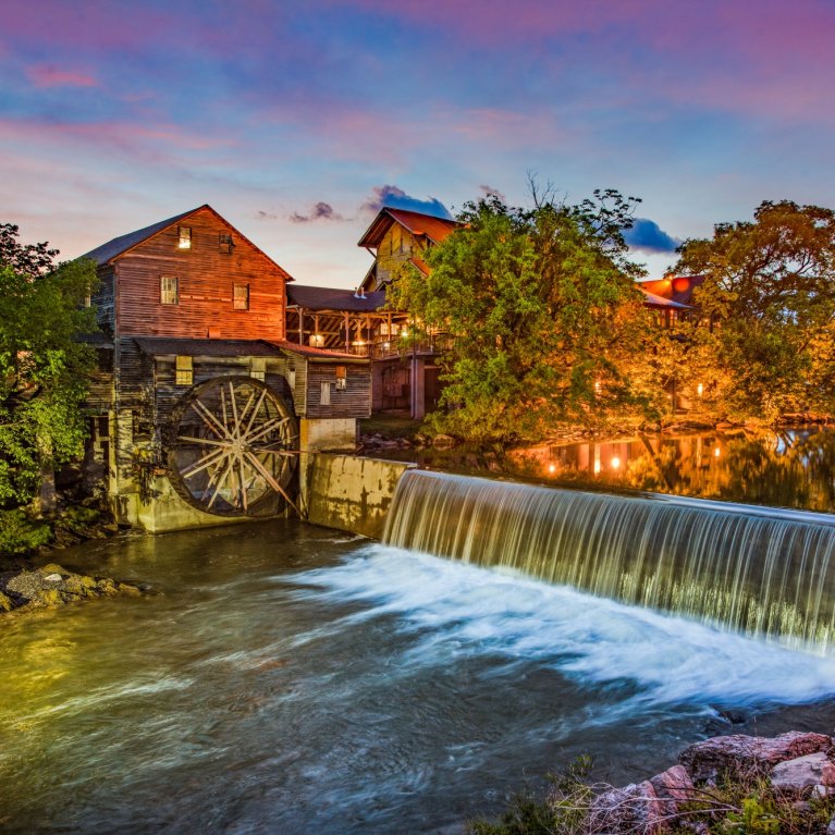 Old mill wheel turns next to waterfall, surroundings illuminated by golden lights, colorful twilight.