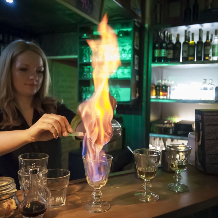 Une femme flambe une boisson derrière le comptoir du bar, tandis que les verres et les bouteilles sont éclairés en arrière-plan.