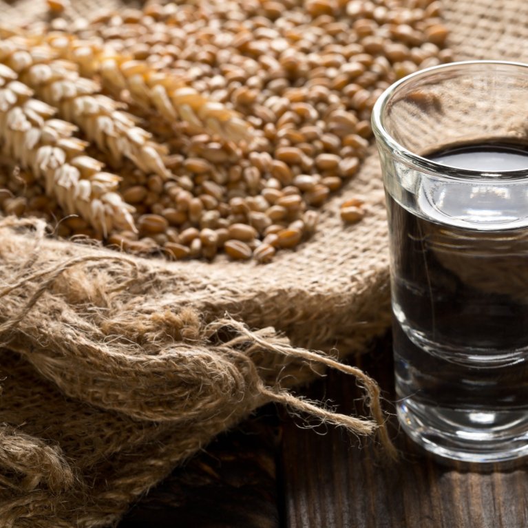 Un verre d'eau-de-vie claire est posé à côté d'épis de céréales sur un sac de jute grossier.