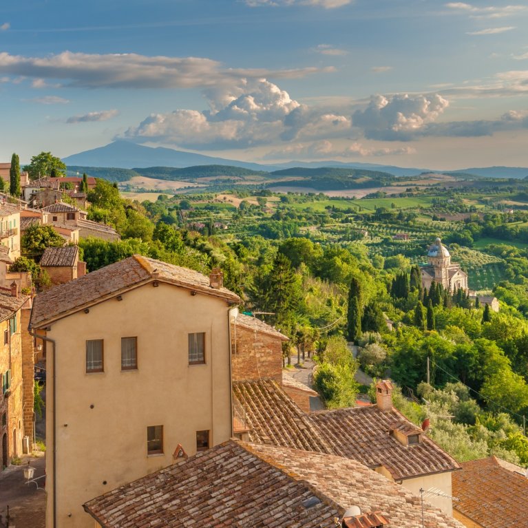 Houses stand sunlit on a hill, surrounded by a lush, green landscape and a wide sky.