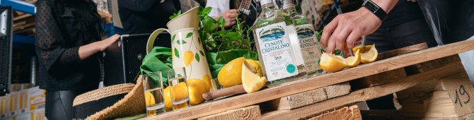 Bottles, lemons and glasses decorate the table; people celebrate happily in the warehouse with guitar music.