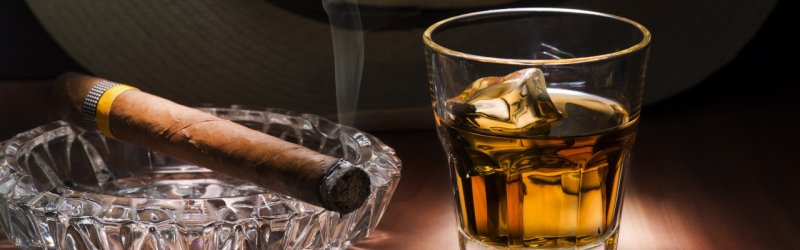 Cigar smoking in ashtray next to whisky glass with ice on wooden table in dim light.