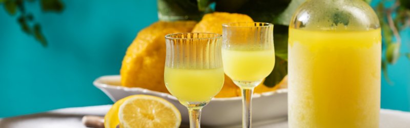 Two glasses of limoncello are placed on a table in front of lemons and a carafe.