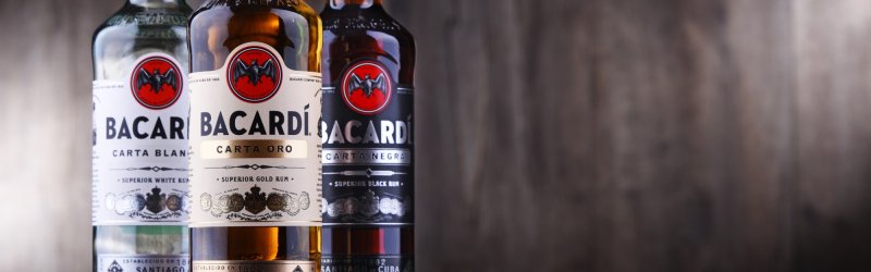 Drie flessen rum staan naast elkaar tegen een vage, houten achtergrond: Bacardi Carta Blanca, Oro, Negra.