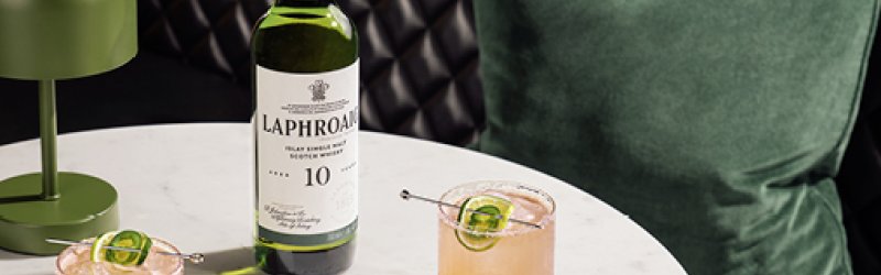 Eine Laphroaig-Whiskeyflasche steht auf rundem Tisch, umgeben von Cocktails und einer Olivenschale.