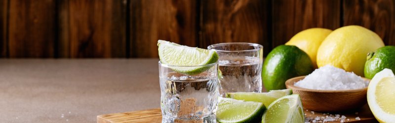 A glass with a slice of lime stands on a wooden board, surrounded by salt and lemons.