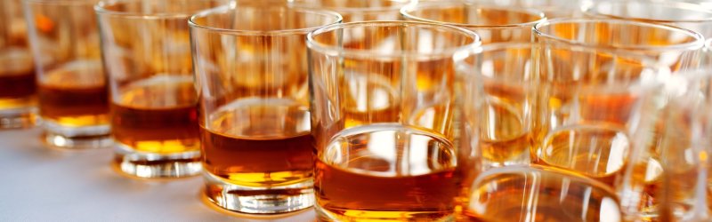 Glasses of amber-colored liquid stand close together on a white surface.