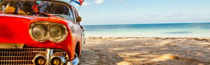 Red vintage car parked on a sandy beach, Cuban flag flying, sea in the background.