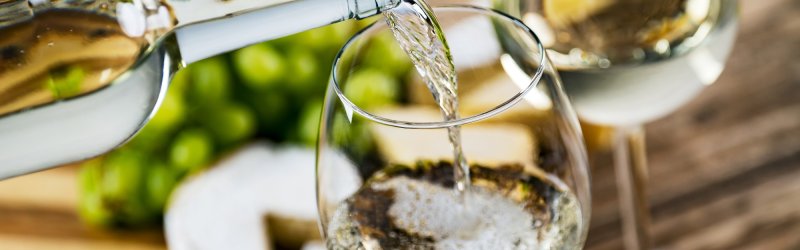 Weißwein wird in Glas eingeschenkt, begleitet von Trauben und Käse auf Holztisch.
