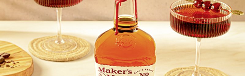 Fles Maker's Mark staat op tafel, geflankeerd door twee cocktailglazen met rode drankjes.