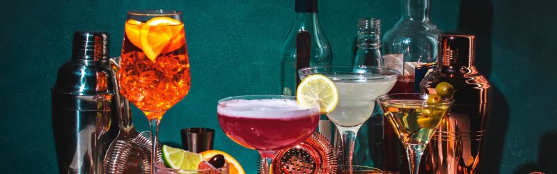 Verschiedene Cocktails stehen auf einem Tisch, umgeben von Shakern und Obststücken.