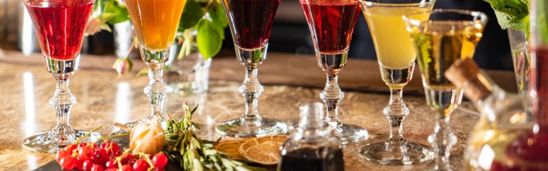 Six colorful cocktails stand on a wooden counter, surrounded by herbs and fruit.
