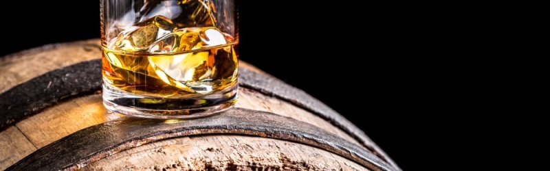 A whisky glass with ice cubes stands on a rustic wooden barrel against a black background.