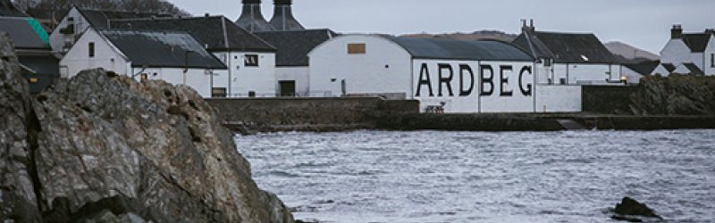 Des bâtiments blancs avec des tours se dressent sur la rive, le nom "ARDBEG" est visible en grand.