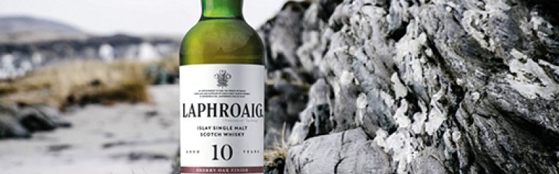 A whisky bottle stands on a rock with a full glass next to it in a natural setting.