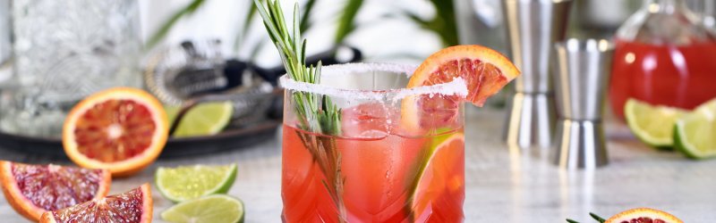 Een glas rode cocktail met een takje rozemarijn, omringd door bloedsinaasappels en schijfjes limoen op een houten tafel.