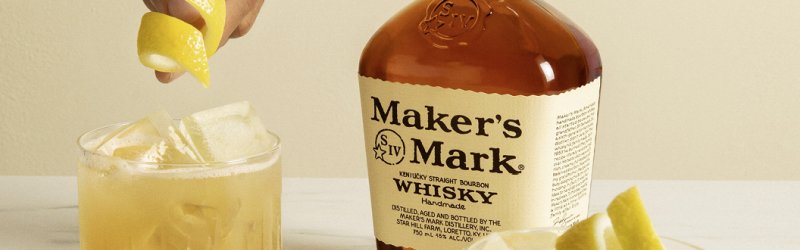 Citroen wordt uitgeperst in een whiskyglas, fles Maker's Mark whisky ernaast op tafel.