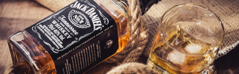 Whiskey bottle lies on a rope, next to it a filled glass with ice cubes on a wooden table.