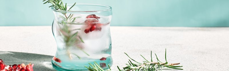 Glas mit Rosmarin und Granatapfelkernen steht auf einem Tisch vor hellblauem Hintergrund.
