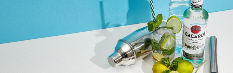 Cocktail ingrediënten gerangschikt op de tafel; Bacardi fles, limoenen, ijsthee, shaker, munt, blauwe en witte achtergrond.