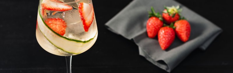 Glass with ice cubes, strawberries and cucumber slices, on a dark table next to strawberries on a napkin.