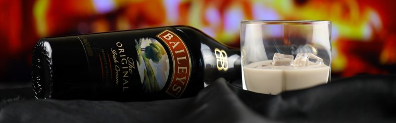 Flasche Baileys liegt neben Glas mit Eiswürfeln auf schwarzem Tuch, bunte Hintergrundlichter.