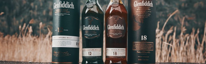 Les bouteilles et canettes de Glenfiddich sont posées sur un sol rocailleux devant un décor de collines boisées.