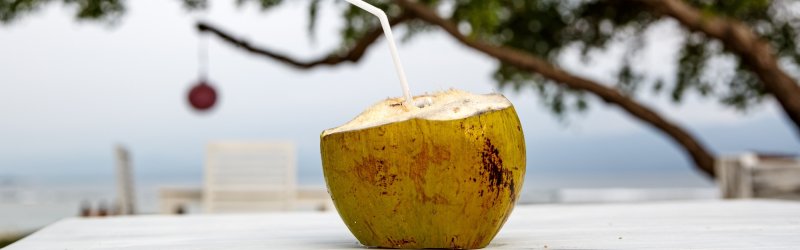 Coconut with straw stands on table, surrounded by beach and blurred trees.