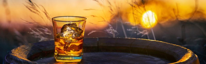 A glass of whisky stands on a barrel with ice cubes, with the sun setting in the background.