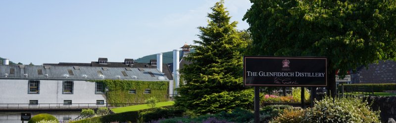Glenfiddich Distillery sign stands in the green garden in front of industrial buildings.