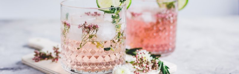 Two pink cocktails with lime slices and flowers are placed on a light-colored board.