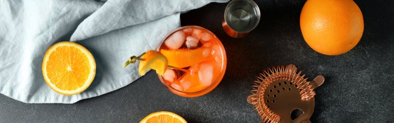 Un verre à cocktail rempli de glace est posé à côté de moitiés d'orange, d'un verre doseur et d'une passoire de bar.
