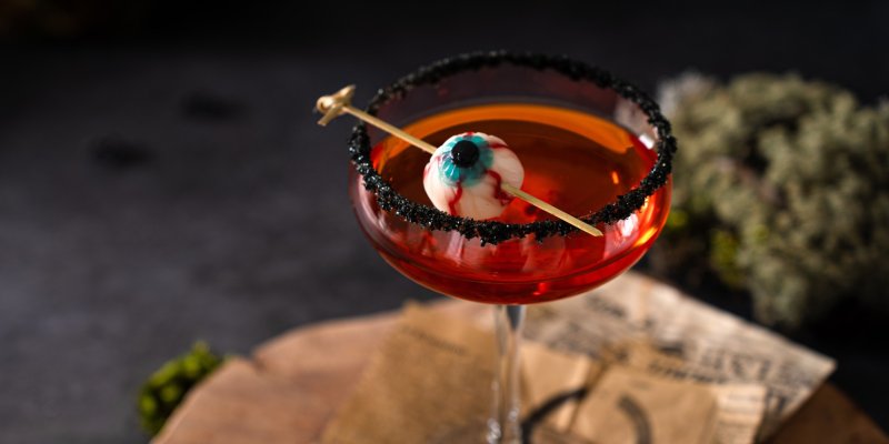 Rotes Cocktailglas, verziert mit "Augen"-Garnitur und schwarzem Zucker, steht auf rustikalem Tisch.