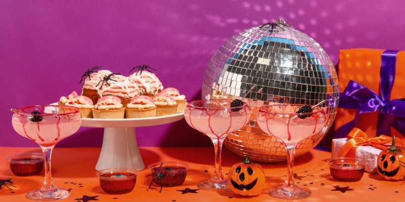 Cocktails stehen dekoriert mit Spinnen und Kürbissen auf einem Tisch vor lila Hintergrund.