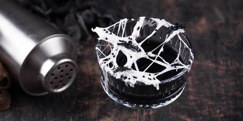 Glas mit Getränk, dekoriert mit Spinnennetz und Spinnen, steht auf dunklem Holztisch.