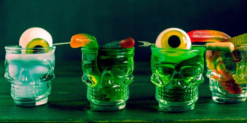 Schädelförmige Gläser enthalten farbige Cocktails mit Augapfel- und Wurmdekorationen auf Holztisch.